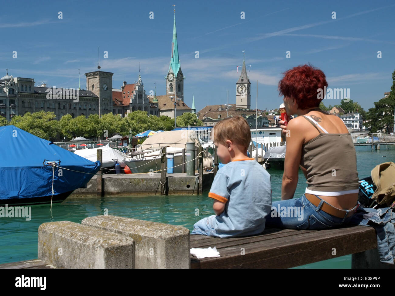 Vista sulla Limmat di Fraumuenster e San Pietro Chiesa; una madre con il figlio seduto su una panchina in primo piano, Switzerl Foto Stock