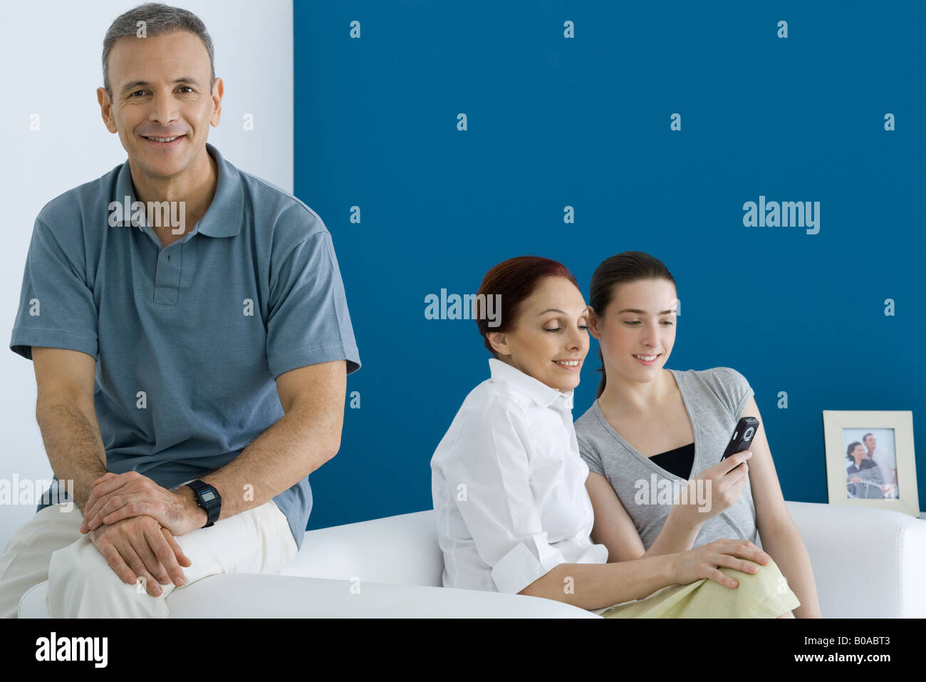 Uomo seduto sul divano, sorridente alla fotocamera, sua moglie e sua figlia guardando il telefono cellulare dietro di lui Foto Stock