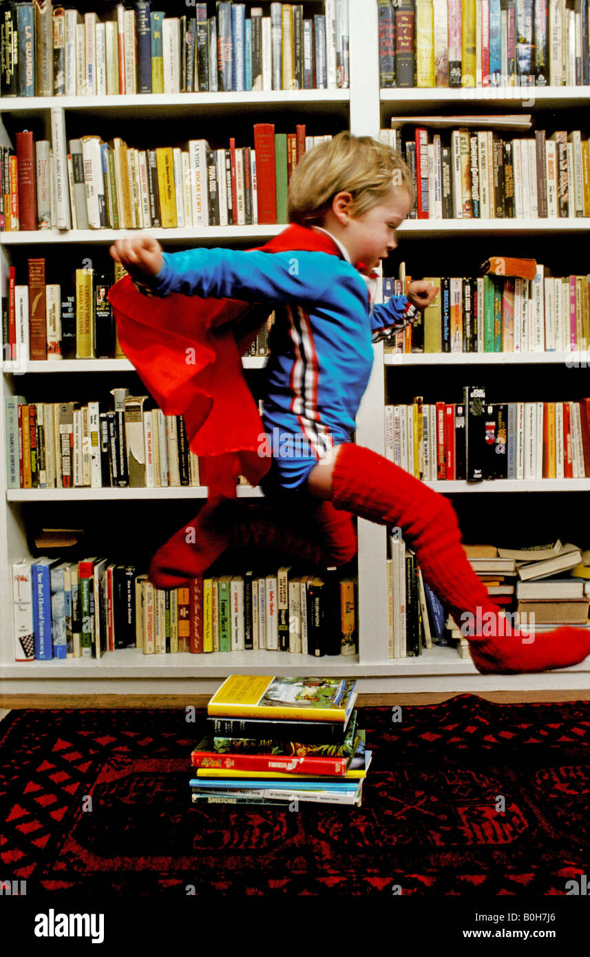 Un bambino di tre anni ragazzo che indossa un capo e gambali corrispondenti salti su una pila di libri come egli pretende di essere Superman Foto Stock