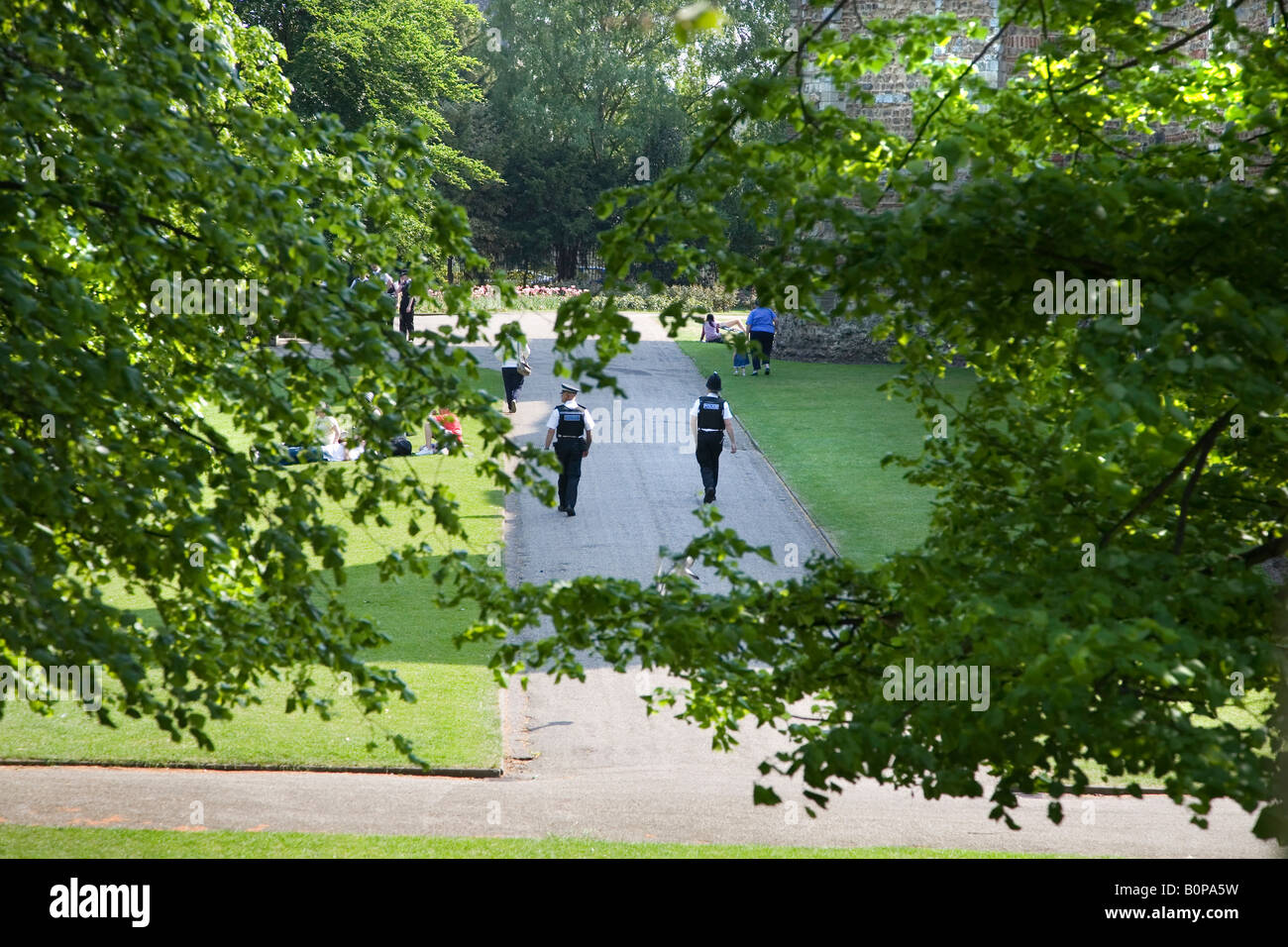Un poliziotto e un poliziotto di pattuglia il COLCHESTER Castle Park in una giornata di sole, visto ATTRAVERSO GLI ALBERI E FOGLIE Foto Stock