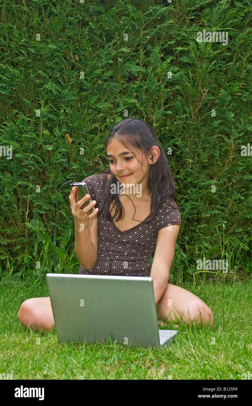 Una giovane ragazza (10 anni) in giardino con il suo computer lap top guardando il suo lettore musicale mp3 con cuffie. Foto Stock