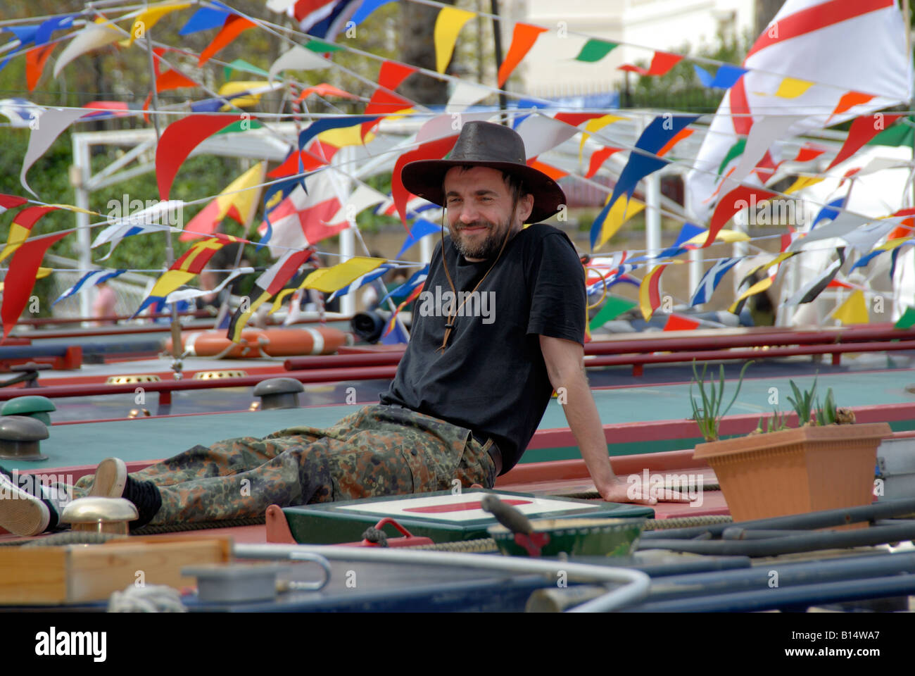 Uomo in cappello da cowboy rilassante sulla sua narrowboat circondato da bandiere e bandierine al Canalway Cavalcata, Little Venice, Londra Foto Stock