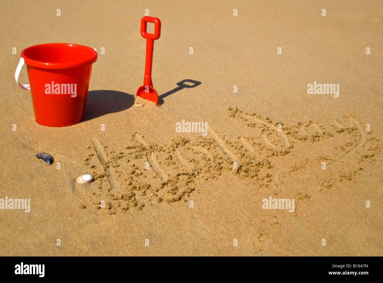 Vista orizzontale di un rosso secchio di plastica e vanga sulla spiaggia accanto a "vacanza" scritto nella sabbia Foto Stock