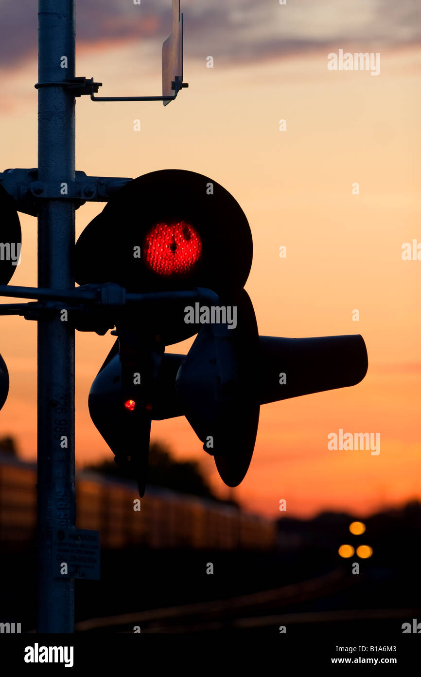 Un treno si avvicina un lampeggiante attraversando il dispositivo di segnalazione come il sole tramonta. Foto Stock