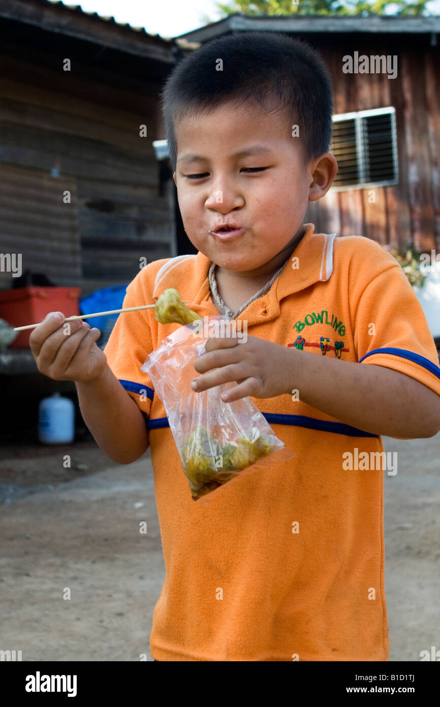 Un piccolo ragazzo tailandese mangia uno spuntino che ha appena comprato da un prodotto alimentare venditore ambulante Foto Stock