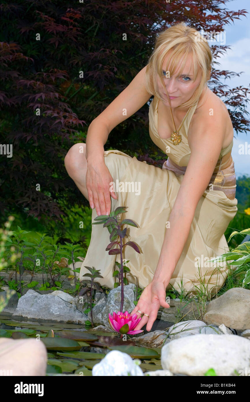 Sposa nel bel vestito d'oro dal laghetto in giardino e che raggiunge la rosa giglio d'acqua. Foto Stock
