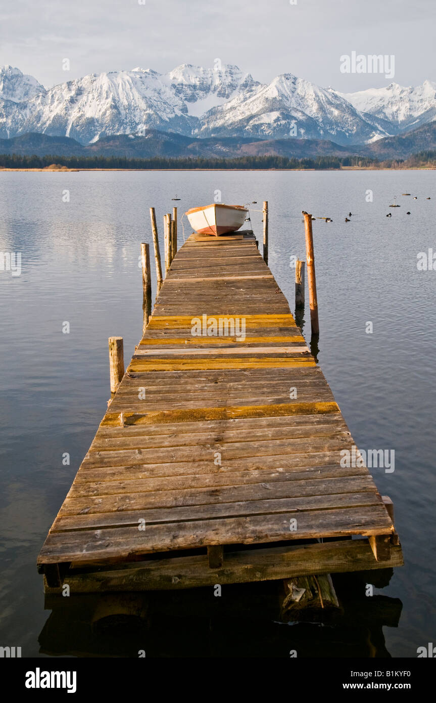 Piccolo canotto sul dock di legno in Hopfensee con montagne coperte di neve in background Baviera Germania Foto Stock