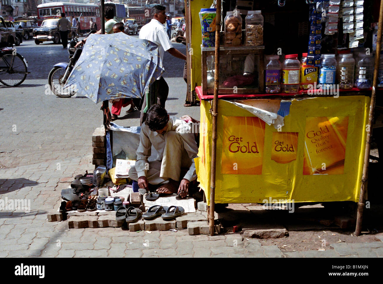 INDIA MUMBAI MAHARASHTRA calzatura repairman imposta il suo negozio su un marciapiede occupato sotto un ombrello come egli mende flip flop a Mumbai Foto Stock