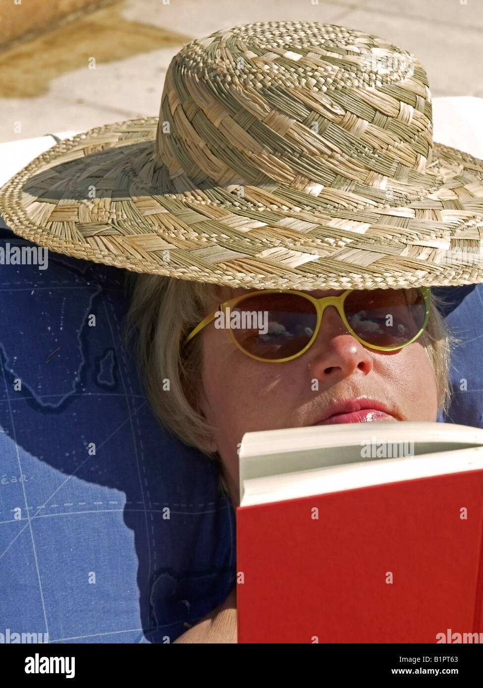 Donna di mezza età con cappello per il sole sdraiati al sole sul lettino la lettura di un libro Foto Stock