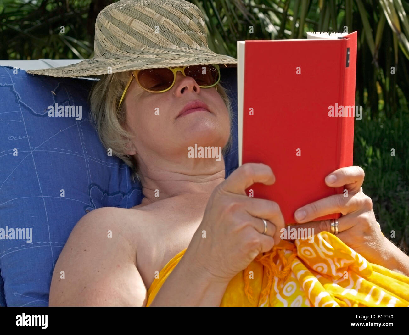 Donna di mezza età con cappello per il sole sdraiati al sole sul lettino la lettura di un libro Foto Stock