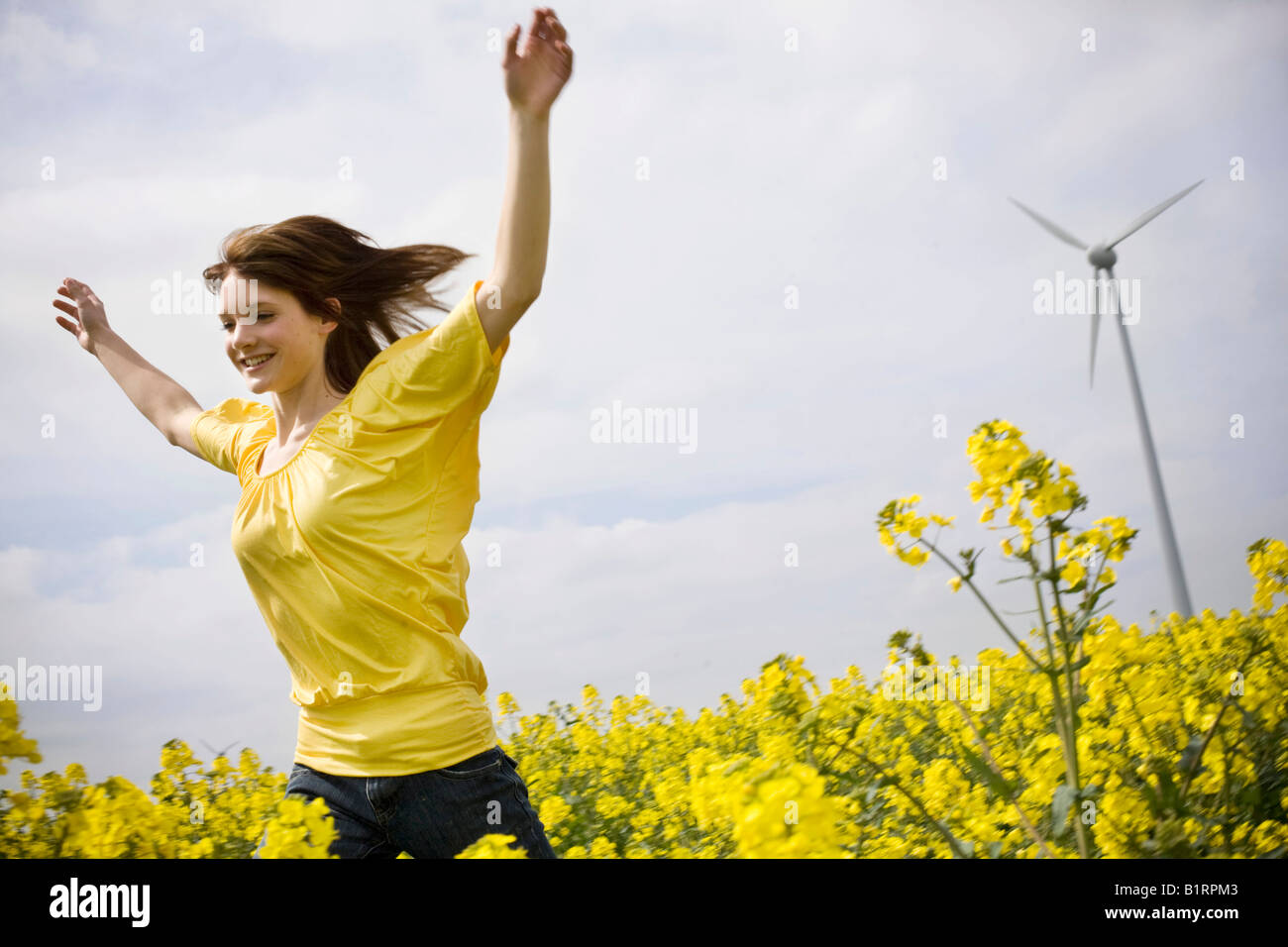 Giovane donna in esecuzione attraverso un canola o campo di colza, turbina eolica nel retro Foto Stock