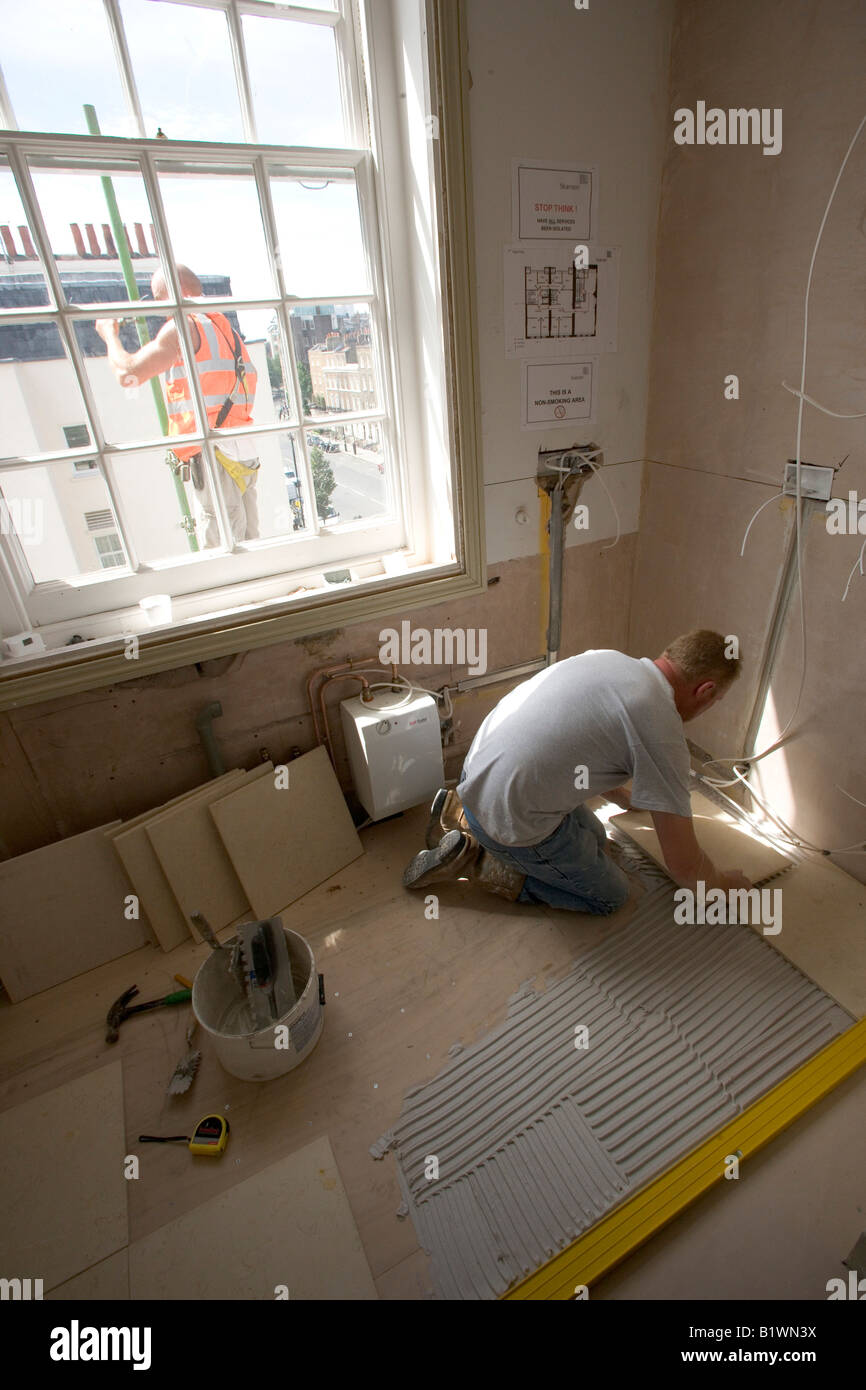 Un lavoratore stabilisce le piastrelle del pavimento per una cucina mentre un ponteggio è essendo costruito all'esterno. Foto Stock