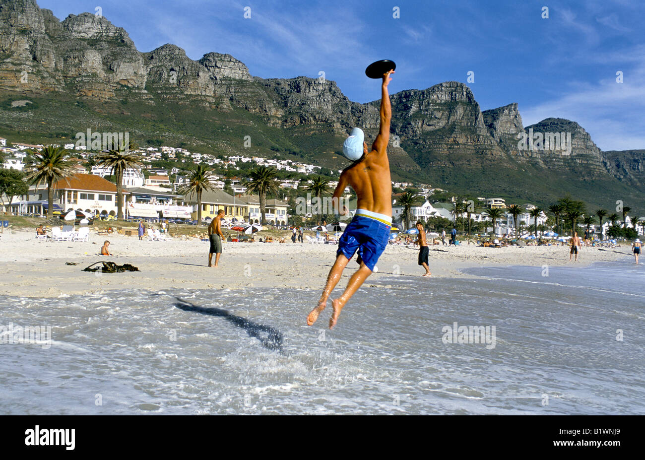 Un uomo salta per la cattura di un frisbee sulla spiaggia di Camps Bay con i dodici apostoli mountain range in distanza. Foto Stock