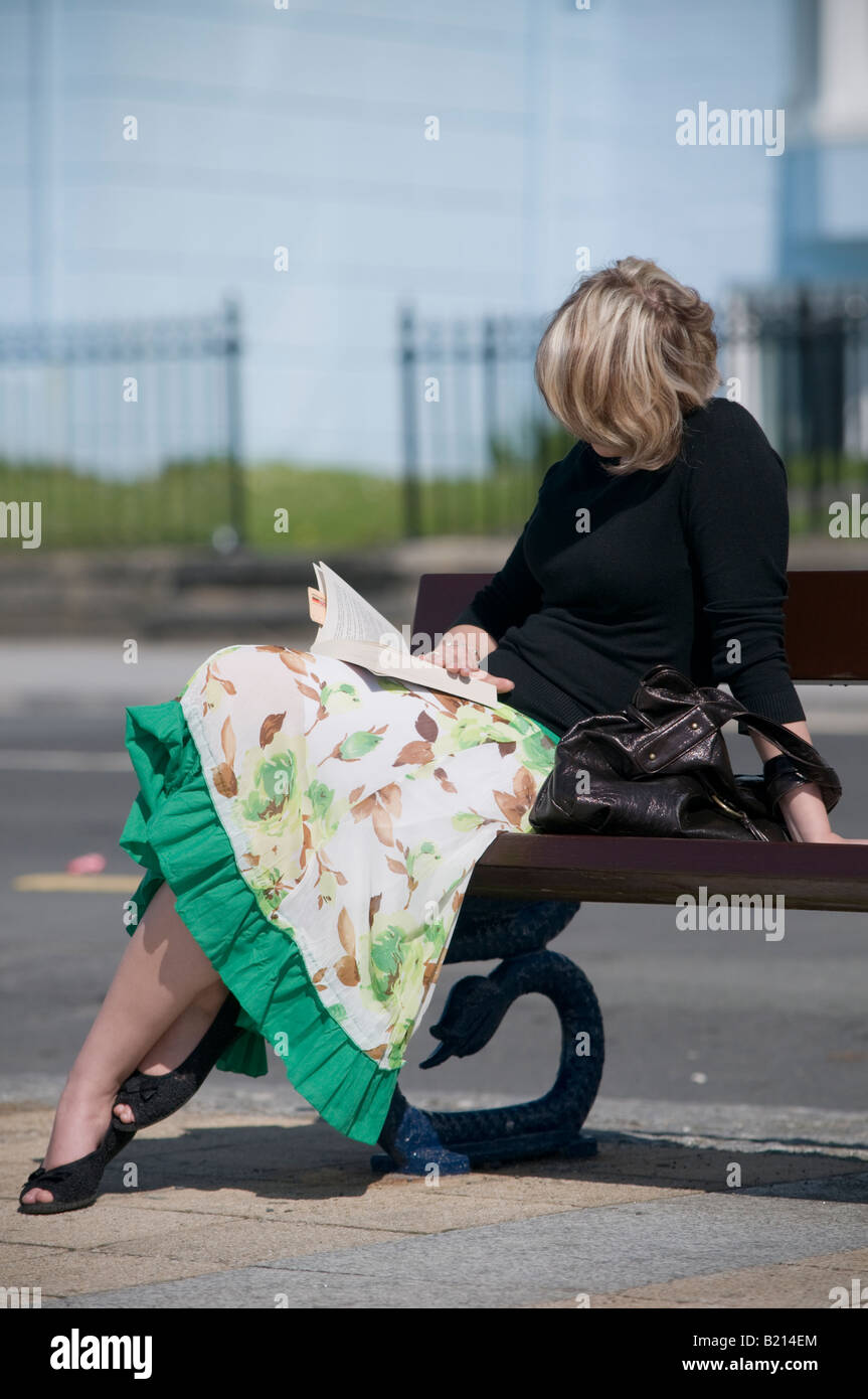 Donna che indossa abiti la lettura di libro in brossura seduta su una panchina in un pomeriggio soleggiato all'aperto Foto Stock