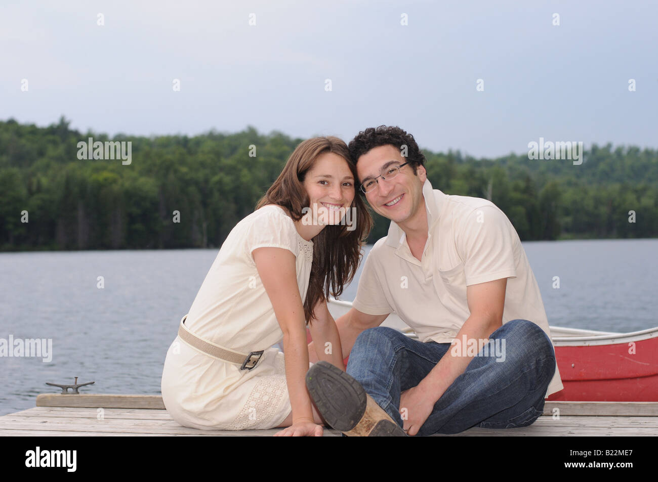 Un felice Caucasian coppia sposata seduto su una dock dall'acqua, con un rosso canoa in background. Foto Stock