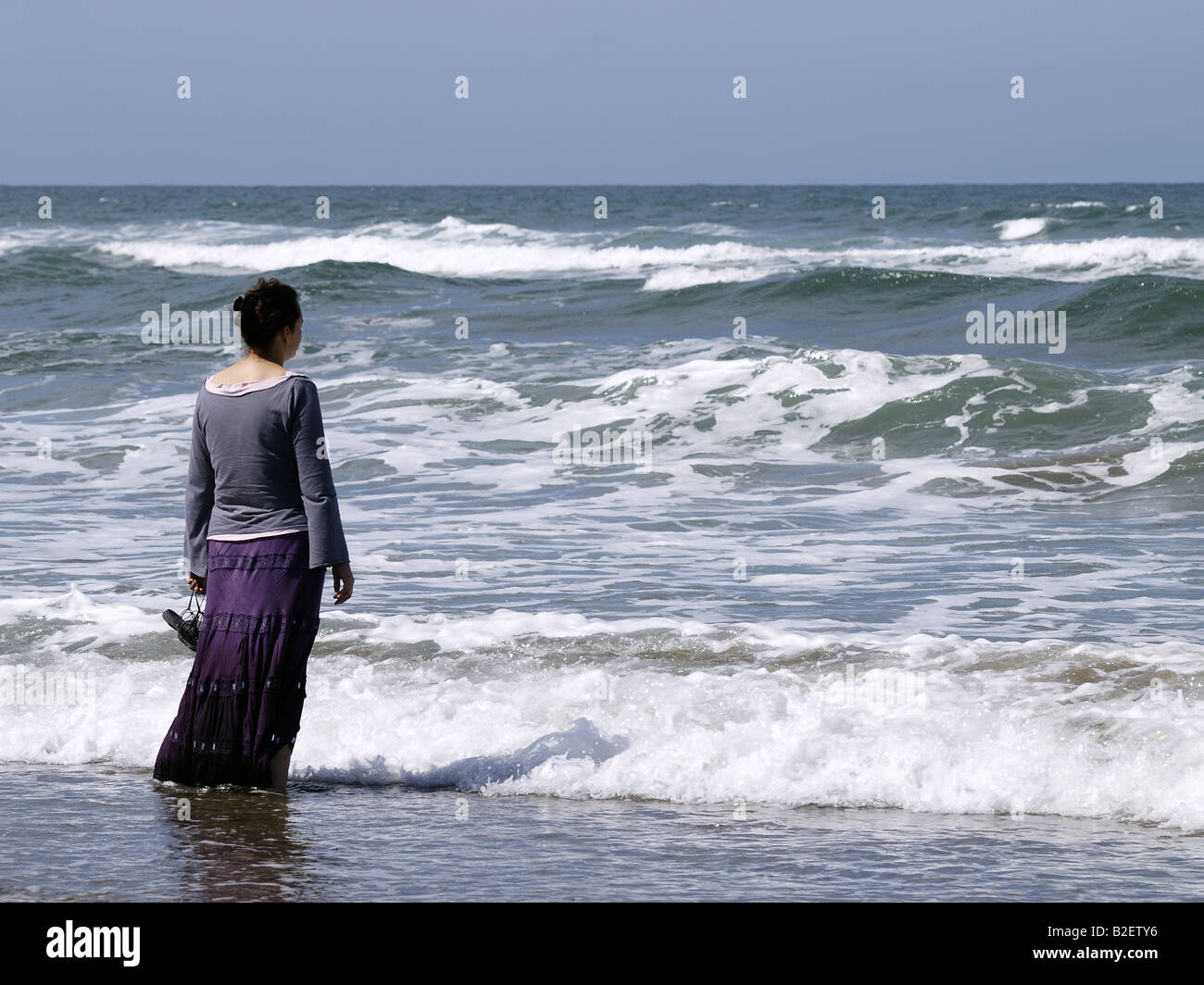 Donna in piedi nel mare, tenendo le sue scarpe con il fondo del suo mantello nell'acqua. Foto Stock