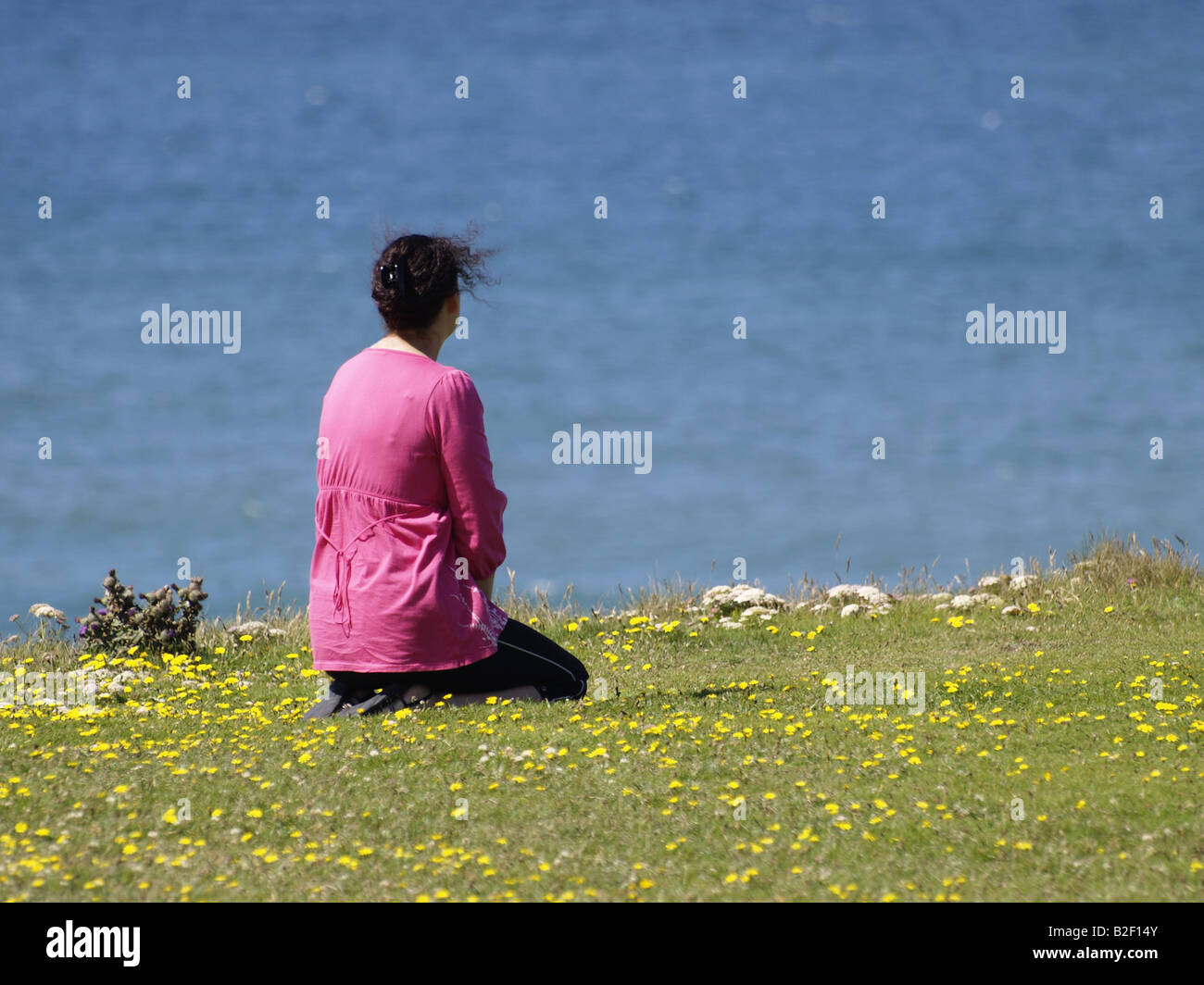 30s donna che indossa un top rosa seduto sulle sue ginocchia, in cima a una scogliera che guarda al mare. Foto Stock