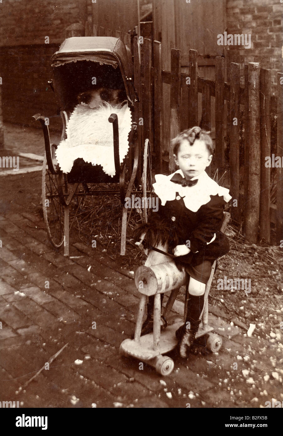 1905 Italiano seppia edoardiana Fotografia di un bambino di 3-4 anni seduto su un trolley tipo cavallo hobby in giardino. PRAM in background. Foto Stock