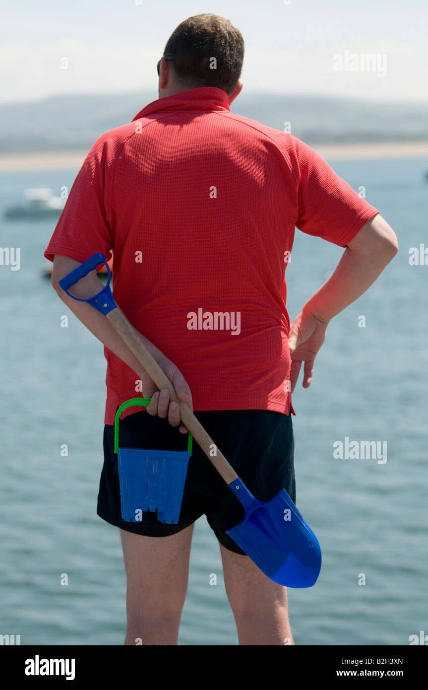 Uomo che indossa camicia rossa azienda secchio di plastica e vanga dietro il suo dorso vacanza estiva, Wales UK visto da dietro Foto Stock