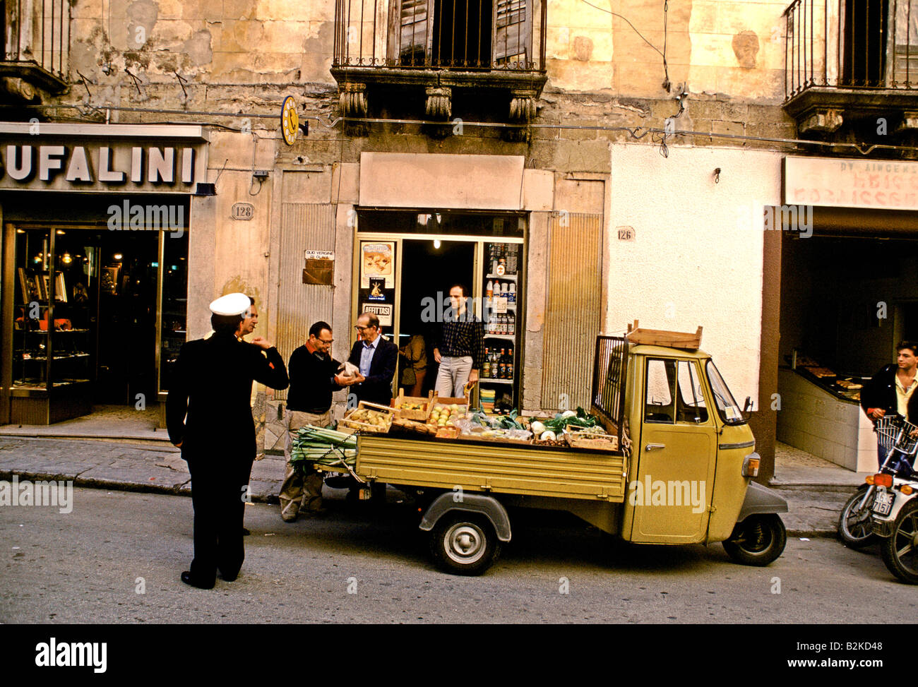Un carrello con frutta fresca e verdura si ferma al di fuori di un negozio 4 uomini e un uomo di polizia come parte di una scena di strada a SCIACCA Foto Stock