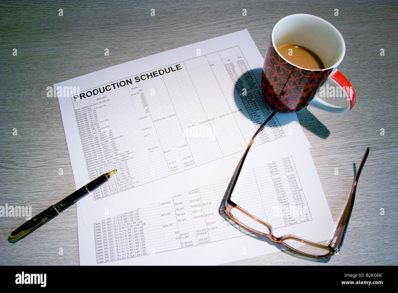 Due fogli di calcolo su una scrivania che mostra un programma di produzione, con una tazza di caffè , una penna e un paio di occhiali Foto Stock