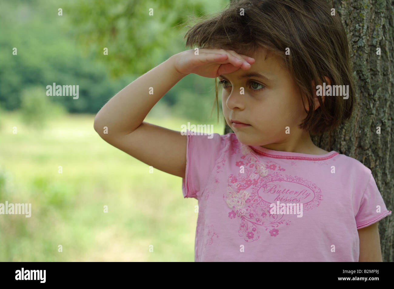 5 anni ragazza sotto l'albero tenendo il suo palm sul suo forehadn cercando di individuare qualcosa di lontano Foto Stock