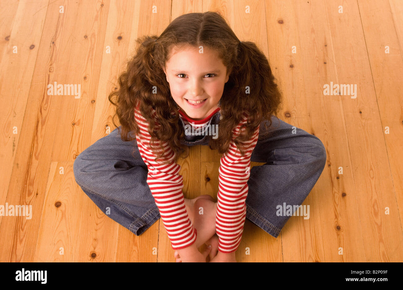 Scatto elevato di una giovane ragazza caucasica con lunghi capelli legati a mazzi, seduta a gambe incrociate su un pavimento di legno, guardando in su sorridendo alla macchina fotografica. Foto Stock