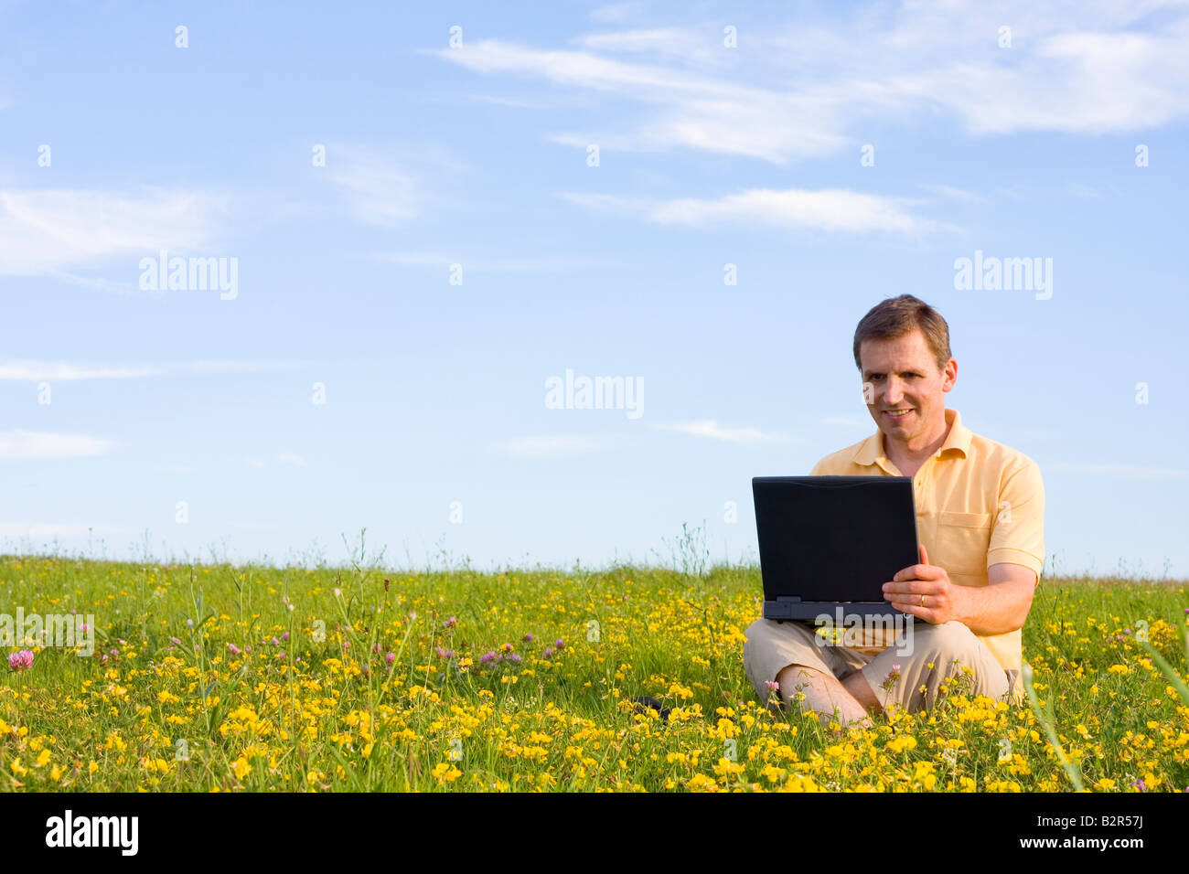 Uomo sorridente seduto con computer portatile in un prato con fiori di colore giallo Foto Stock