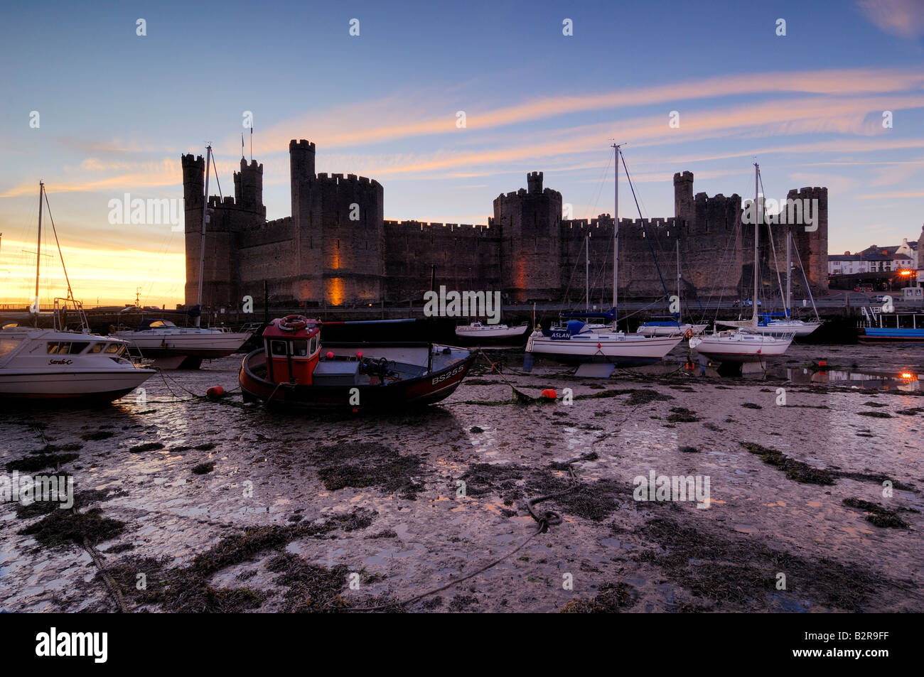 Caernarfon Castle sulla costa del Galles del Nord illuminata di notte con le barche in estuario spiaggiata sulla bassa marea Foto Stock