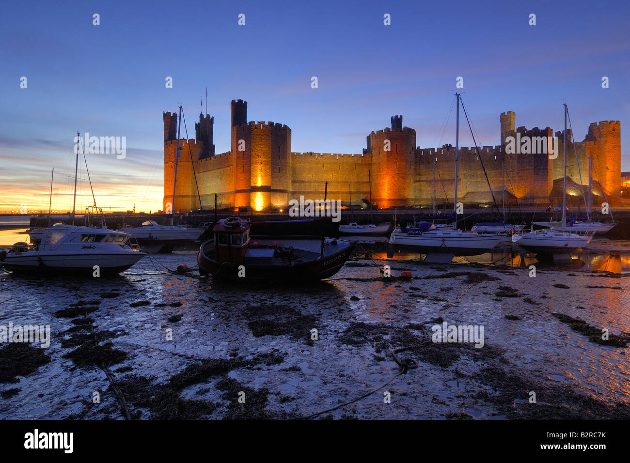 Caernarfon Castle sulla costa del Galles del Nord illuminata di notte con le barche in estuario spiaggiata sulla bassa marea Foto Stock