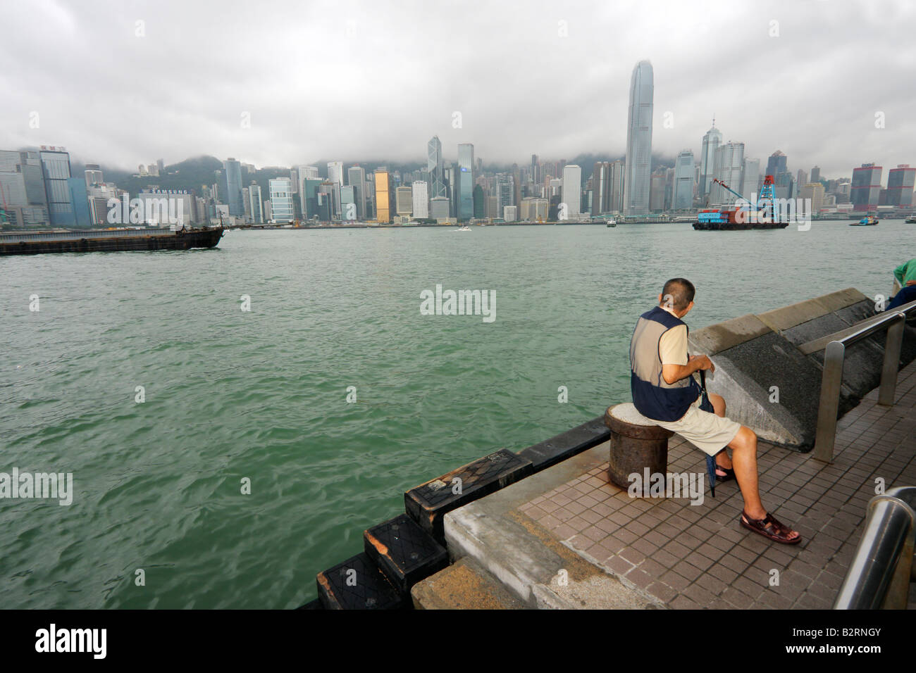 Un uomo cinese seduto sul lato di Kowloon di Victoria Bay cercando di fronte ai grattacieli di Isola di Hong Kong Foto Stock