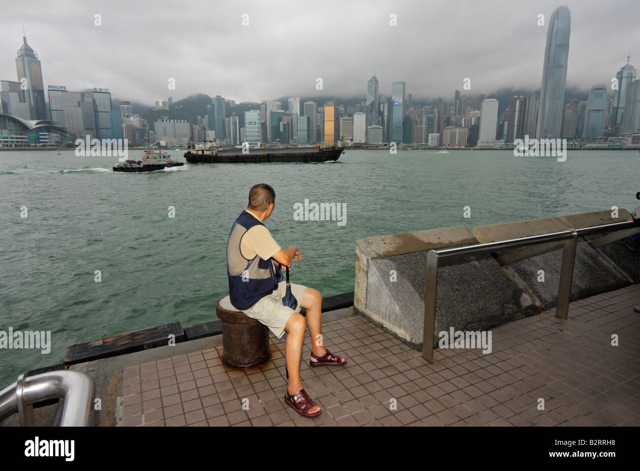 Un uomo cinese seduto sul lato di Kowloon di Victoria Bay cercando di fronte ai grattacieli di Isola di Hong Kong Foto Stock