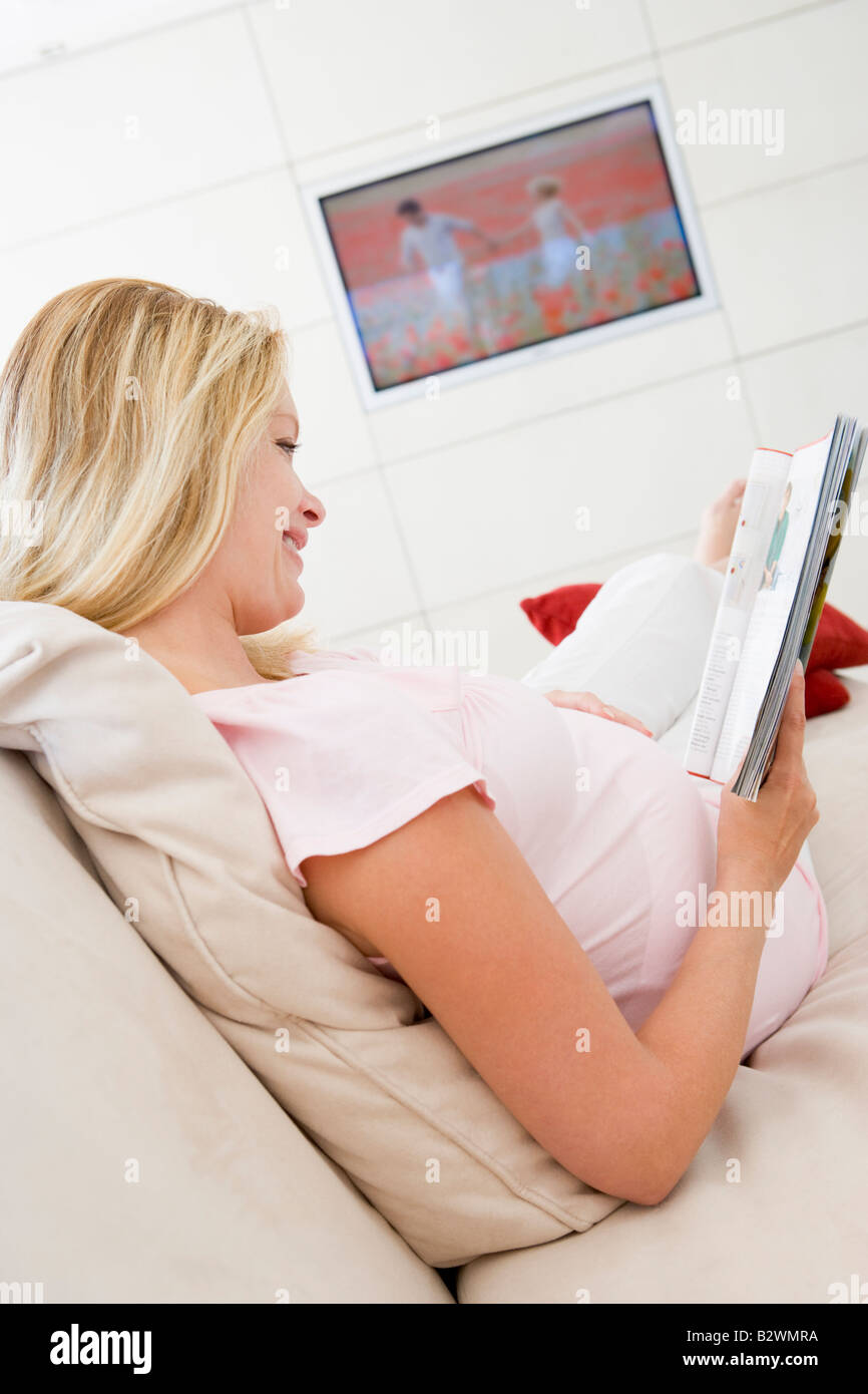 Donna incinta rivista di lettura con televisione in background sorridente Foto Stock
