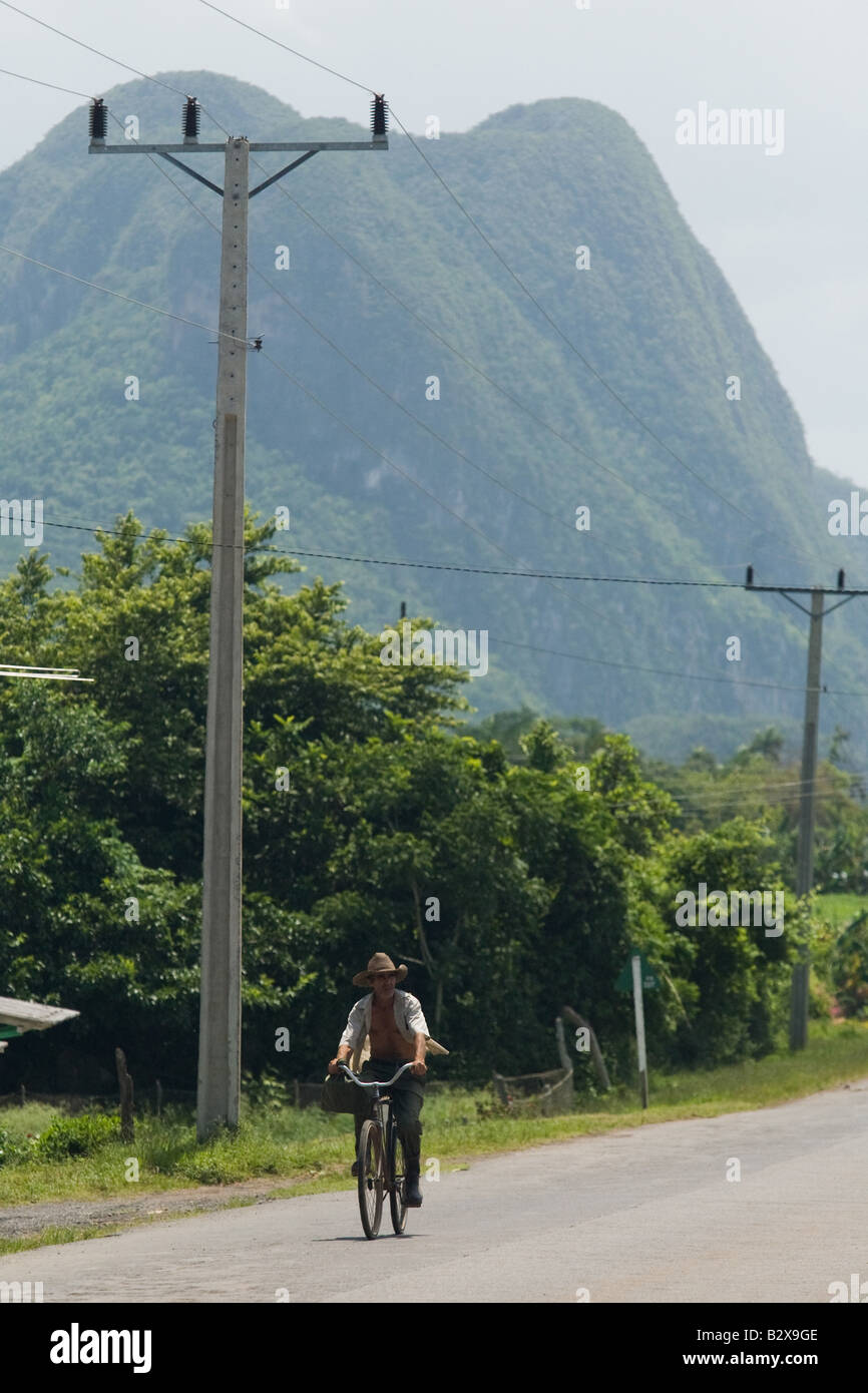 Un uomo che cavalca la sua bicicletta lungo una strada dominato da una grande formazione di montagna chiamato mogote vicino a Vinales Cuba Foto Stock