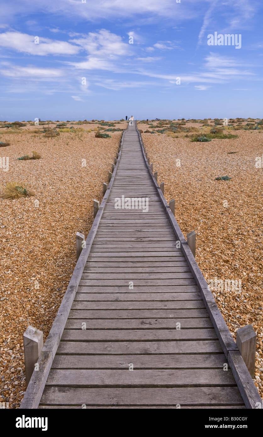 Passerella in legno si estende nella distanza lungo la spiaggia di ciottoli di Dungeness nel Kent in una giornata di sole Foto Stock