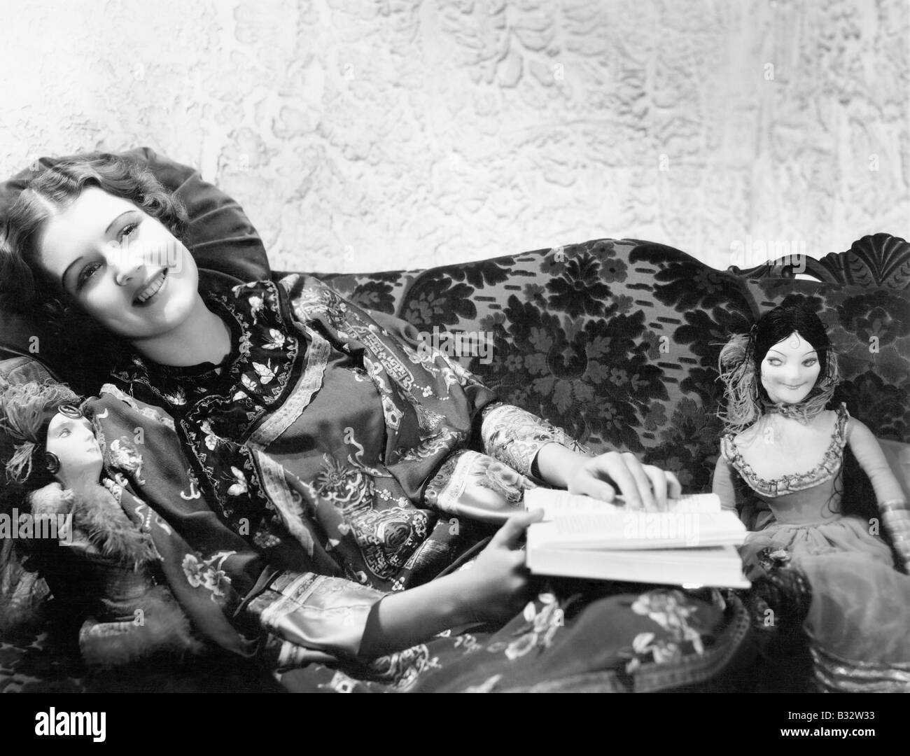 La donna in una stampa cinese vestito sdraiato sul divano con le sue bambole e un libro Foto Stock