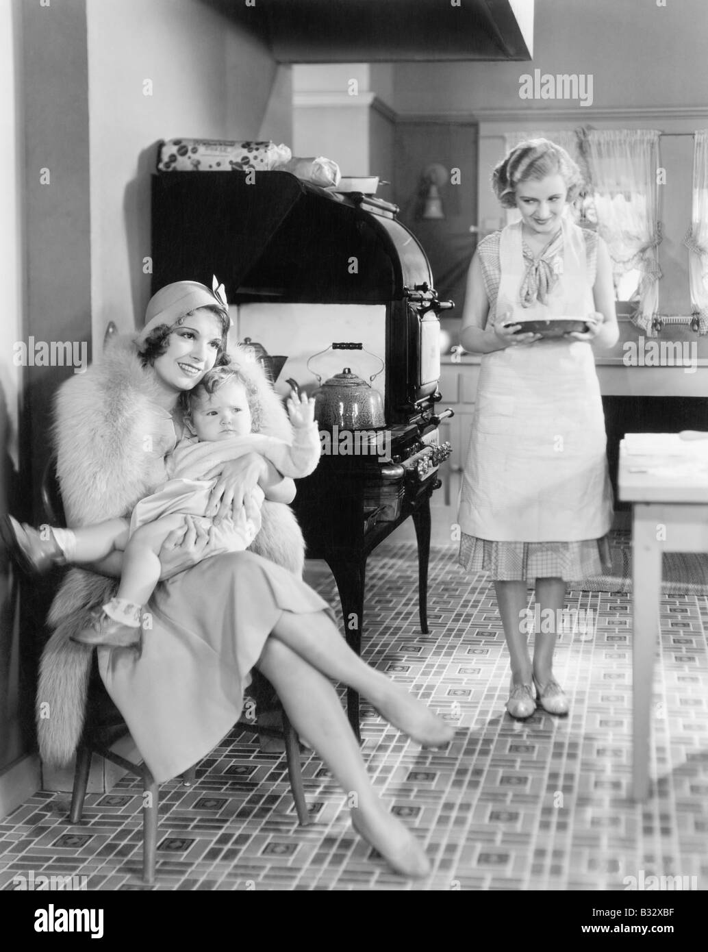 Eleganti Donna seduta con il suo bambino in una cucina mentre un amico sta portando un grafico a torta Foto Stock