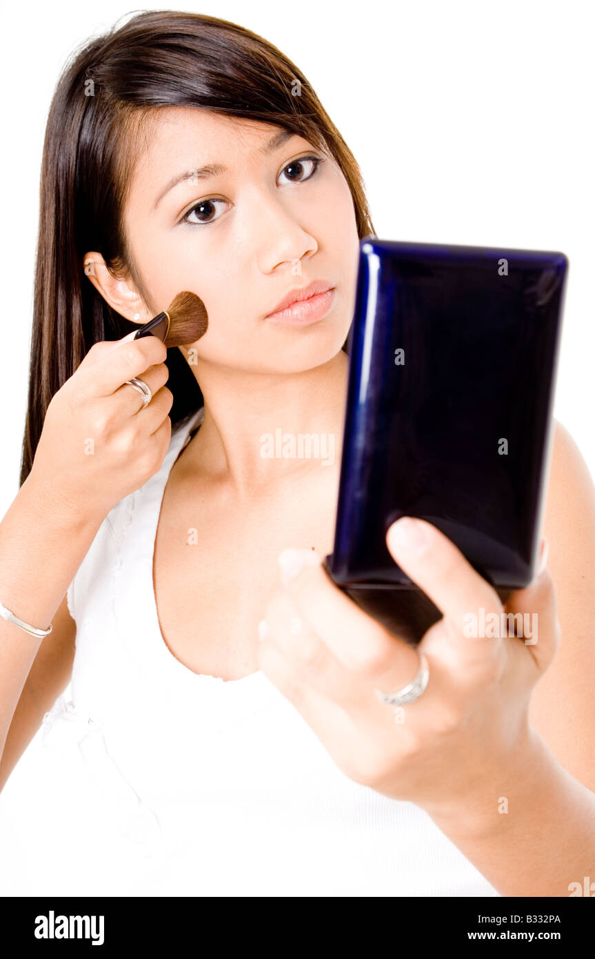 Una bella giovane donna in Asia si applica foundation per la sua pelle con un pennello mentre guardando in uno specchio tascabile (focus sulla donna) Foto Stock