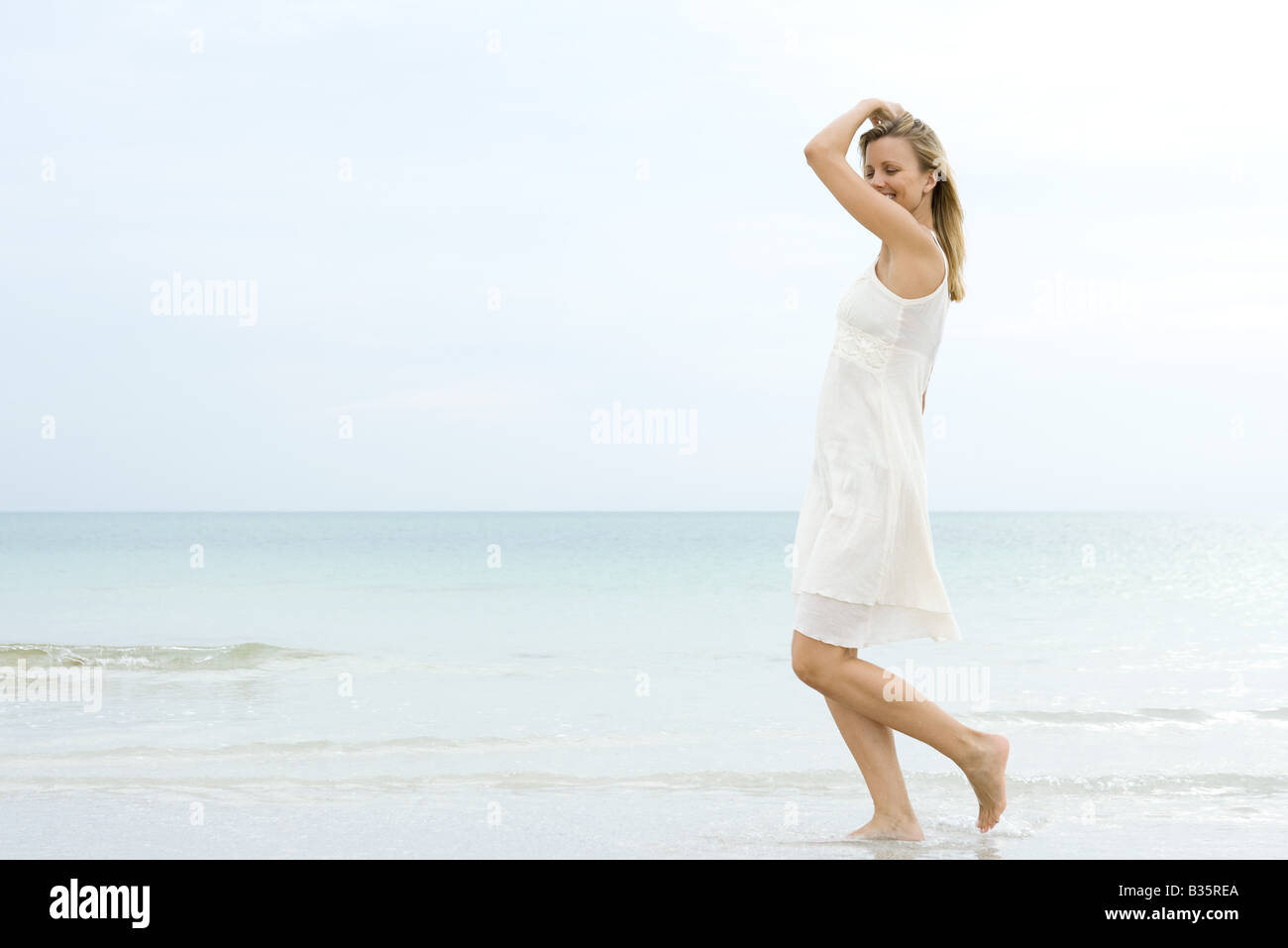 La donna a piedi la spiaggia nel surf, sorridente, vista laterale Foto Stock