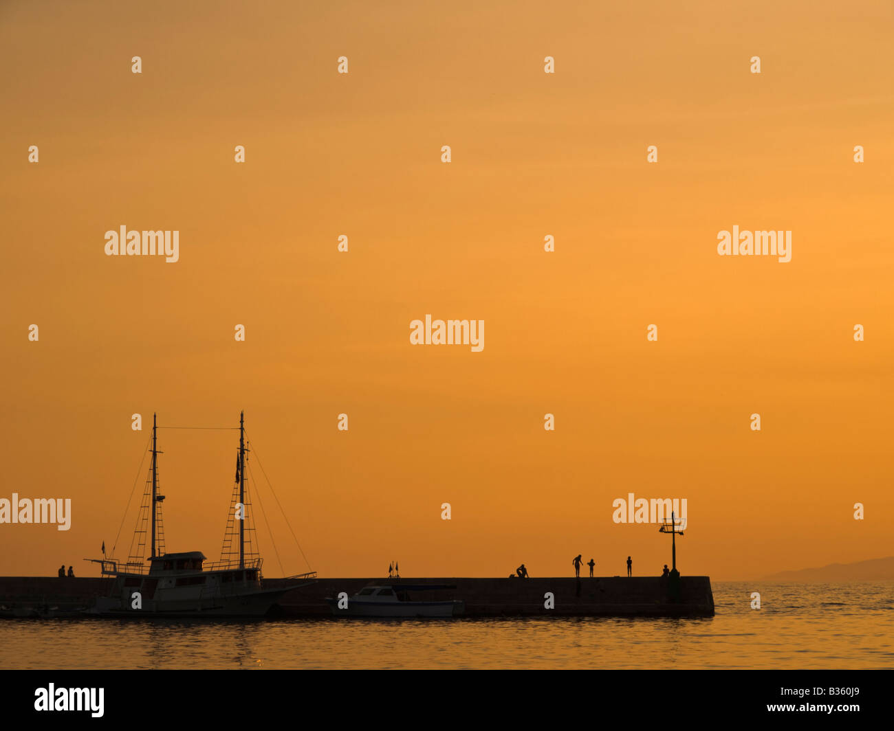 Il tramonto. Otto persone in piedi sul molo del piccolo porto cittadino. Una nave ormeggiata può essere visto. Foto Stock