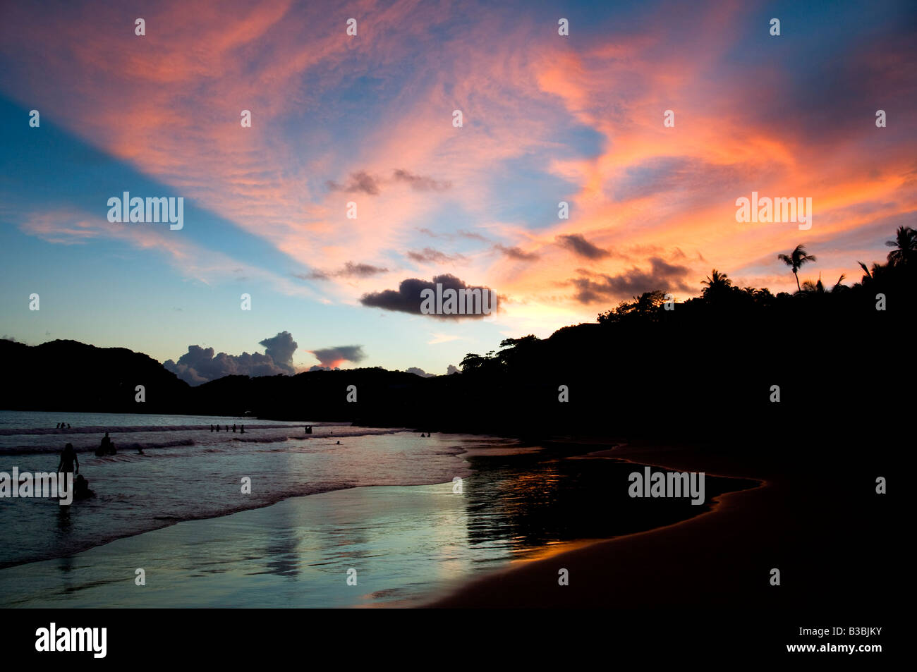Playa la Ropa Beach Sunset Zihuatanejo Messico | ZIHUATANEJO, Guerrero, Messico - figure sagomate si snodano ai margini di Playa la Ropa mentre il cielo sopra il Bahía de Zihuatanejo brucia arancione e rosa al crepuscolo. Incorniciata da promontori scuri e colline coperte di palme, la spiaggia si trova a sud del centro della città ed è conosciuta per l'acqua più tranquilla e riparata di Playa Principal. Foto Stock