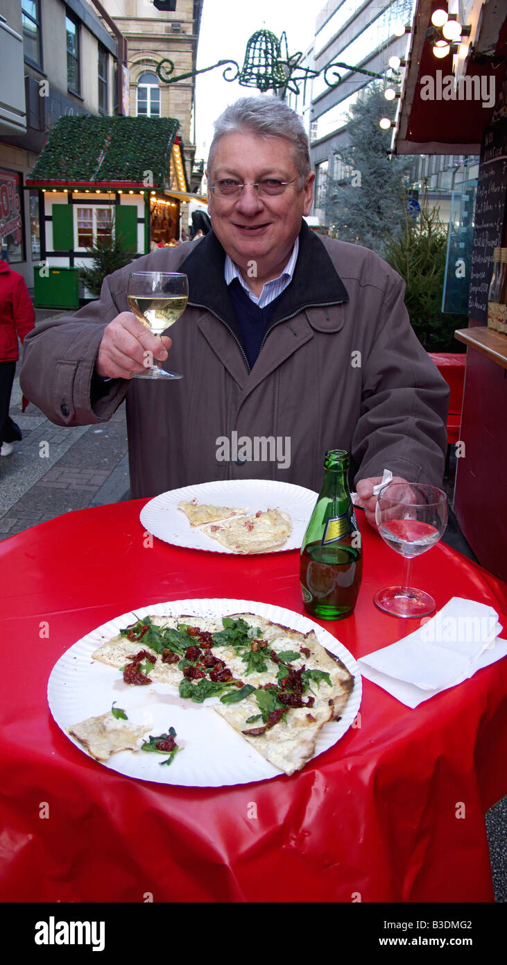Mercatino di Natale di Wuppertal-Elberfeld, alimenti e bevande, l uomo sta a un tavolo bar e un brindisi per il fotografo con un bicchiere di vino bianco, due piastre con Tarte flambee stand sul tavolo Foto Stock