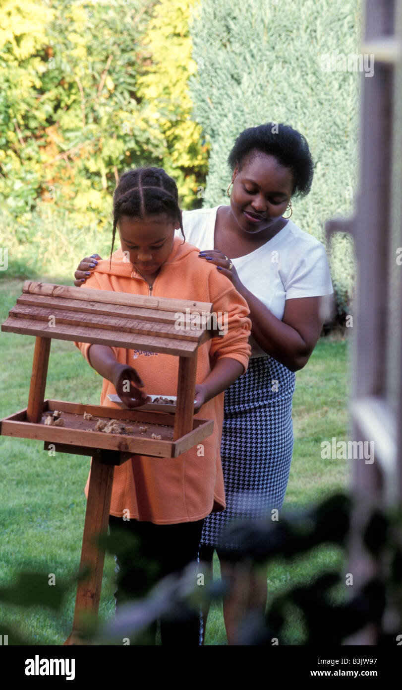 Nero a madre e figlia di mettere il cibo sulla tavola di Uccelli nel giardino cottage Foto Stock