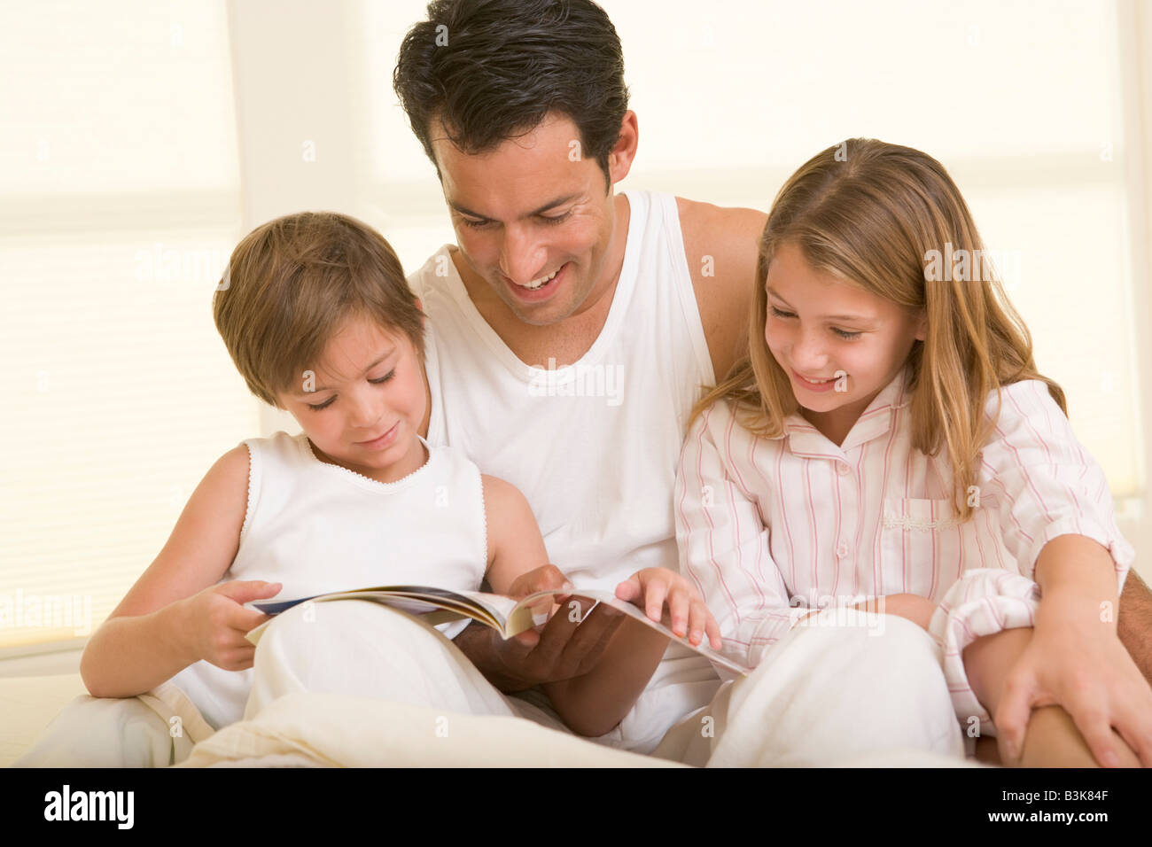 Uomo con due giovani bambini seduti nel letto a leggere un libro e sorridente Foto Stock