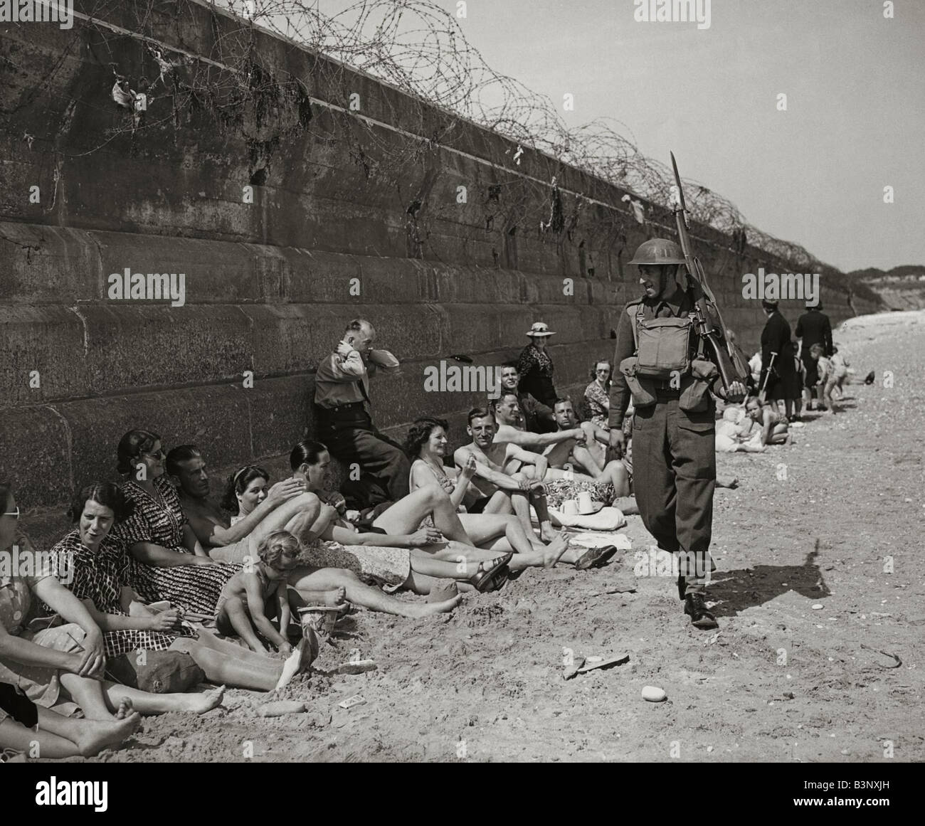 I turisti a prendere il sole pityingly guardare al passaggio di un home veteran anteriore portante un fucile e baionetta stagno che indossa il casco e maschera a gas vacanze WW2 soldato parete mare filo spinato seaside beach Luglio 1941 Foto Stock