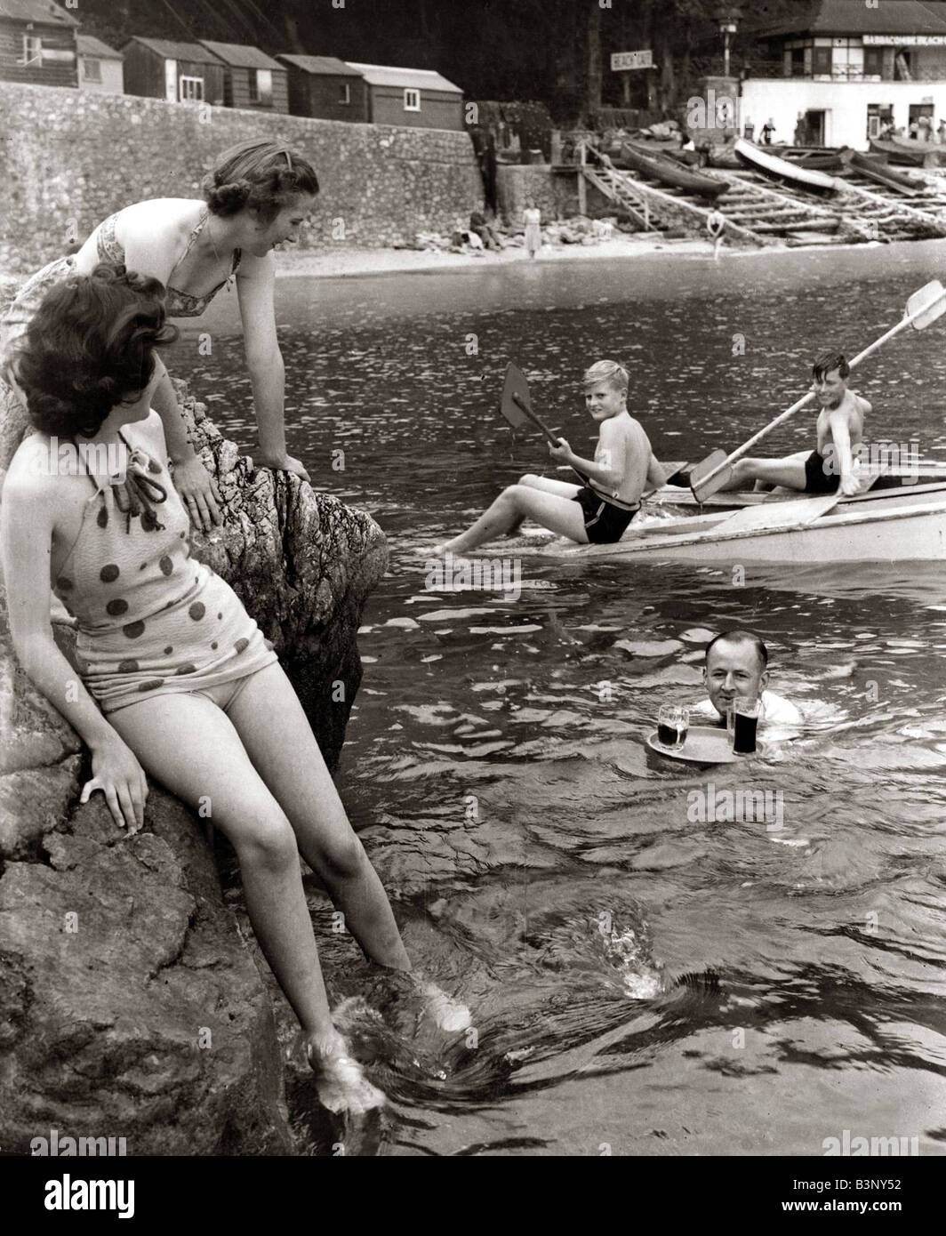 Una famiglia seduta sulle rocce e giocare in acqua a Babbacombe Bay nel Devon come cameriere nuota fuori dal bar a portare sulle bevande per loro Settembre 1948 Foto Stock