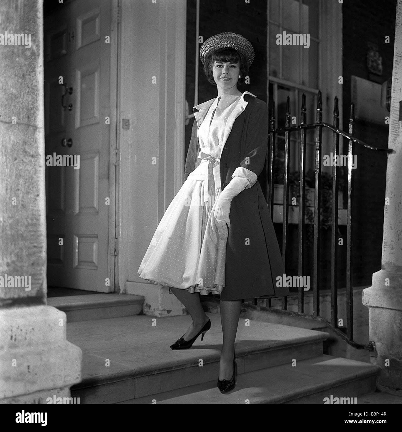 Un modello che indossa un design alla moda nel 1962 indossa un cappotto lungo su un intero costeggiato il vestito in piedi sui gradini di una casa Foto Stock