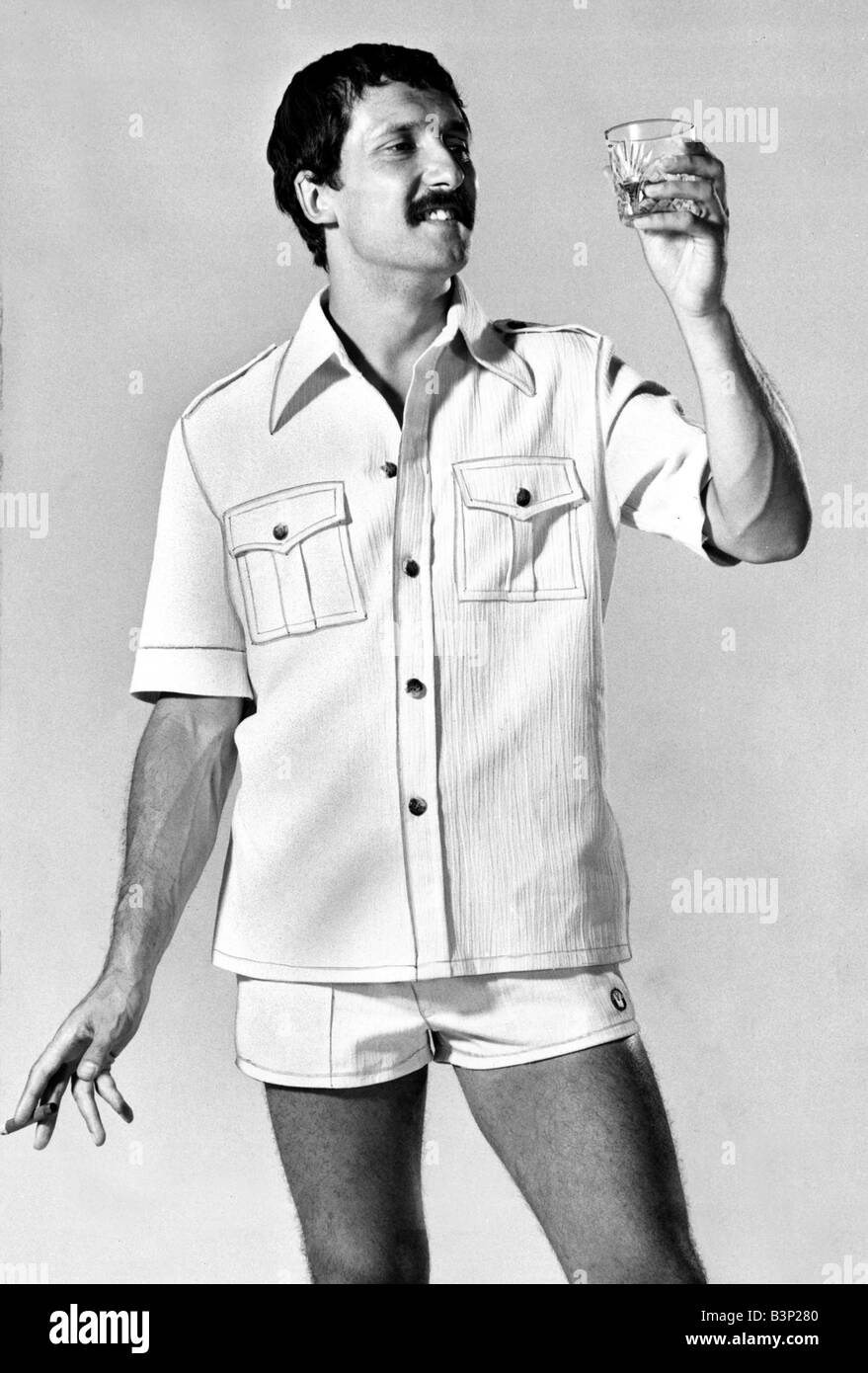 Un uomo che indossa una camicia con pantaloncini guardando un bicchiere di whisky e tenendo un sigaro Agosto 1976 modello maschile mens moda uomini fumatori di bere whisky degli anni settanta sveglia specchio 13 8 76 Foto Stock