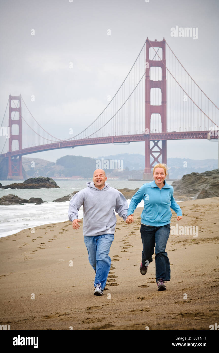 Una giovane coppia tenendo le mani che corre lungo la spiaggia di fronte al Golden Gate Bridge di San Francisco Foto Stock