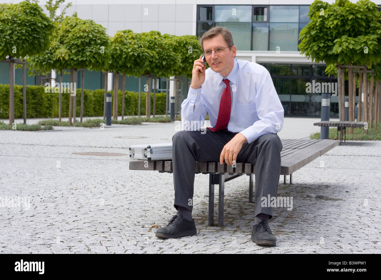 Imprenditore parlando al telefono cellulare mentre è seduto su una panchina davanti a un edificio per uffici Foto Stock
